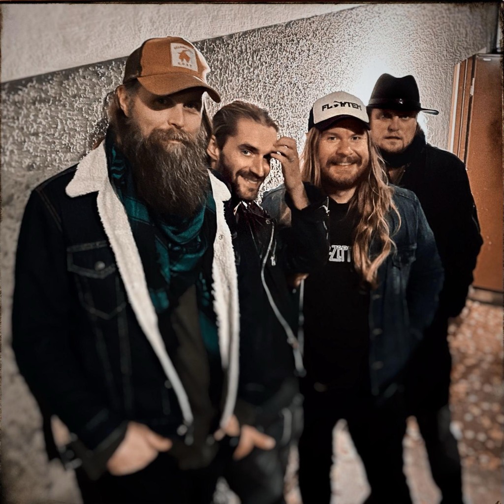 Pressbild på Flykten: fyra bandmedlemmar poserar mot en ljus putsad vägg. En av dem bär en keps med bandnamnet.